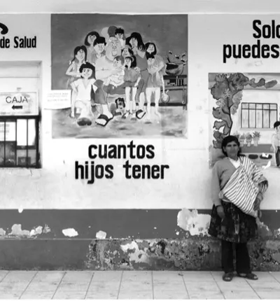 Black-and-white photo of a public health clinic wall with family-planning murals reading ‘cuántos hijos tener’ and ‘Solo tú puedes decidir’; a woman stands at right carrying a baby in a woven sling.