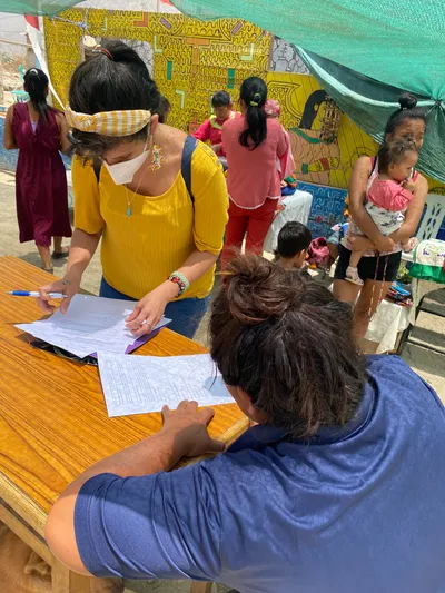 A woman in a yellow shirt signs a document at an outdoor table, with others and a baby visible in the background.