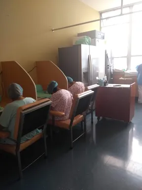 The image shows several individuals with blue medical caps seated in chairs, in what appears to be a room with yellow walls and a few pieces of furniture, including a refrigerator.