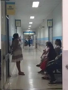A brightly lit hospital hallway with a shiny floor, a clock on the wall, and a sign that reads EsSalud. Several people are seated and waiting, with medical personnel visible further down the hallway.
